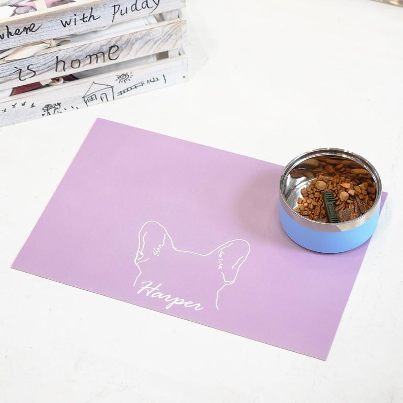 May include: A light purple pet placemat with a white outline of a dog's head and the name "Harper" in script. A stainless steel and light blue pet bowl filled with food sits on the right. A wooden crate with text is in the background.