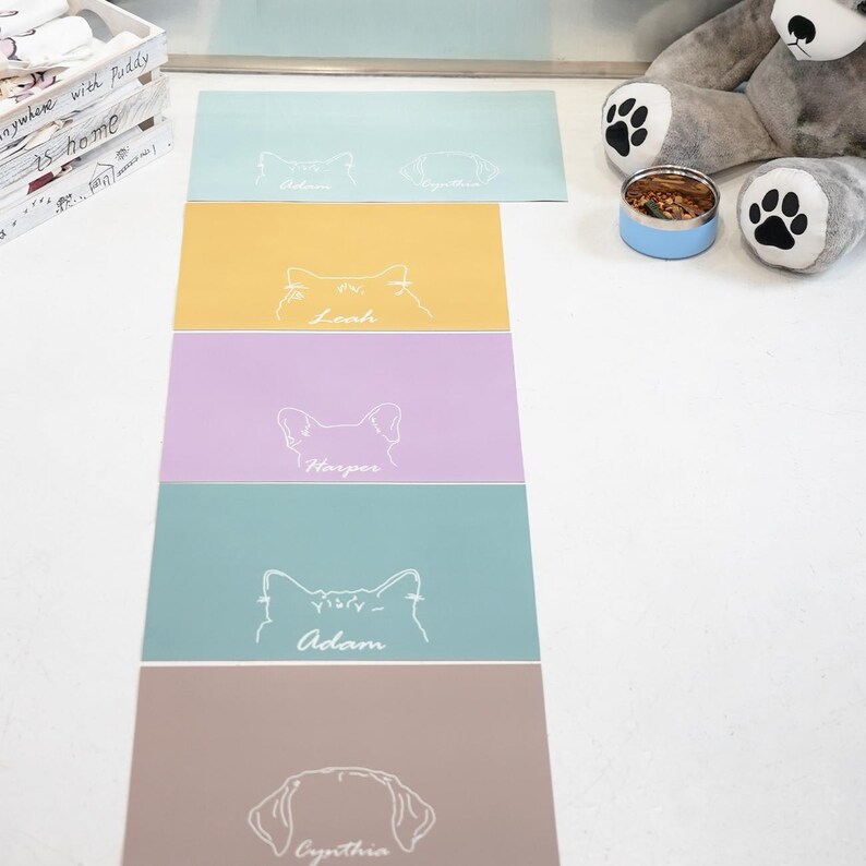May include: A collection of colorful pet placemats. Each placemat features a pet's name and a line drawing of a cat or dog. The placemats are in shades of blue, yellow, purple, and brown.
