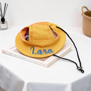 Puede incluir: Sombrero de pescador amarillo con el nombre "Lara" bordado en azul. El sombrero tiene una correa de barbilla negra y está sobre un libro. El color del sombrero es amarillo brillante y soleado.