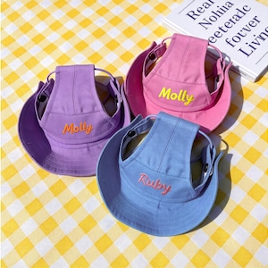 Puede incluir: Tres sombreros para perros en tonos morado, rosa y azul, cada uno con ala ancha y correa. Los sombreros están bordados con los nombres "Molly" y "Ruby" en un color contrastante. Los sombreros están sobre una superficie a cuadros amarillos y blancos.