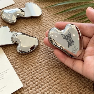 May include: Silver-coloured heart-shaped gua sha tool featuring a dog portrait and the name "Arthur". Other gua sha tools are visible on a textured surface.