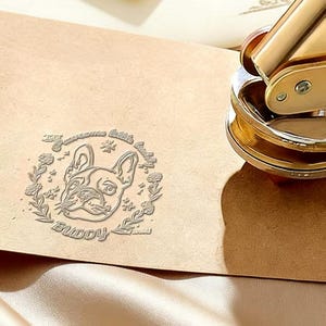 May include: A gold-colored embossing seal next to a tan envelope with a debossed image of a French Bulldog. The image is surrounded by floral elements and the words "Awesome Cuddle Buddy".