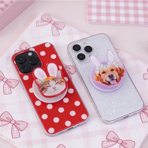 Può includere: Due custodie per telefono con pop socket. Una custodia è rossa a pois bianchi con un gatto con orecchie da coniglio. L'altra custodia è glitterata argento con un cane con orecchie da coniglio. Entrambe le custodie sono su una superficie a fantasia rosa e bianca.