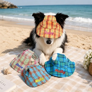 May include: A dog wearing a yellow, red, and orange plaid visor with the name "Ruby" embroidered on it. Three additional plaid visors in green, blue, and orange are nearby, each with a name embroidered on them.