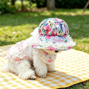 May include: A small white dog dressed in a floral hat, dress, and bandana. The hat features a blue, pink, and white floral design with the name "Stella". The dress and bandana have a matching floral pattern with lace trim. The dog is on a yellow and white checked blanket.