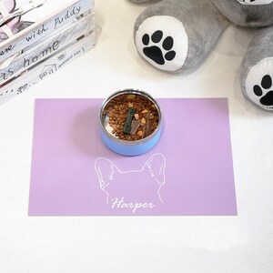 May include: A light blue pet food bowl filled with dry food and treats sits on a lavender placemat. The placemat features a dog silhouette and the name "Harper" in white. A gray stuffed animal with paw prints is in the background.