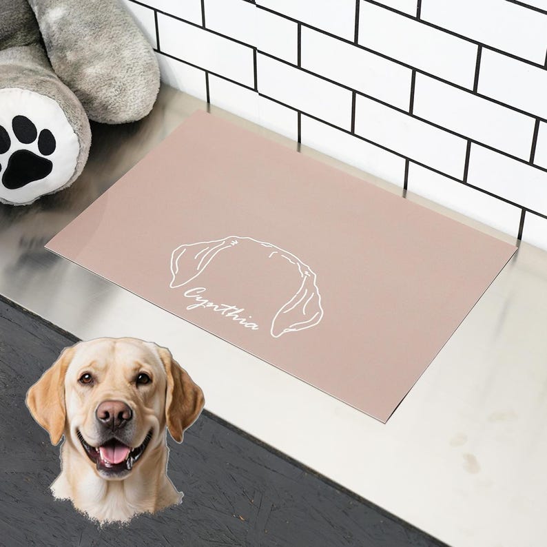 May include: A rectangular, blush-colored doormat with a white outline of a dog's head and the name "Cynthia" in script. A smiling Labrador Retriever is in the lower left corner. The mat is on a metallic surface.