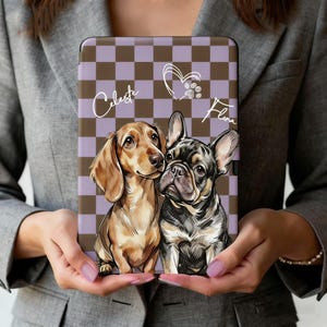 Może przedstawiać: Etui na tablet z ilustracją jamnika i buldoga francuskiego. Tło ma brązowo-fioletowy wzór w kratkę. Imiona "Celeste" i "Flina" są napisane kursywą, z sercem i odciskami łap.