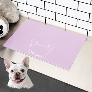 May include: A lavender pet placemat with a white outline of a dog's head and the name "Harper" in script. A white French Bulldog is in the foreground. A gray dog toy is in the upper left corner. The background is a white brick wall.