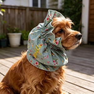Puede incluir: Un gorro para perro de color verde azulado con un estampado floral de flores rosas, rojas y azules. La palabra "Molly" está bordada en amarillo. El gorro está recogido en la parte superior y lo lleva un Cocker Spaniel.