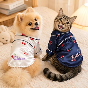 Op de afbeelding: Een hond en een kat dragen bijpassende pyjama's met kersenprint. De pyjama van de hond is wit met de naam "Chloe" in paars geborduurd. De pyjama van de kat is marineblauw met de naam "Jade" in roze geborduurd.