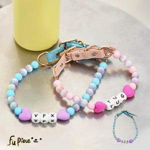 May include: Two dog collars with beaded strands in pastel colours. One collar is light blue, and the other is pink. Each collar has a leather strap and gold-coloured hardware. The collars have heart-shaped beads and lettered beads spelling "VFX" and "TUG".