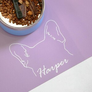 May include: A light purple pet food mat with a white outline of a dog's head and the name "Harper" in cursive. A blue and silver pet food bowl is partially visible, filled with dry food and treats.