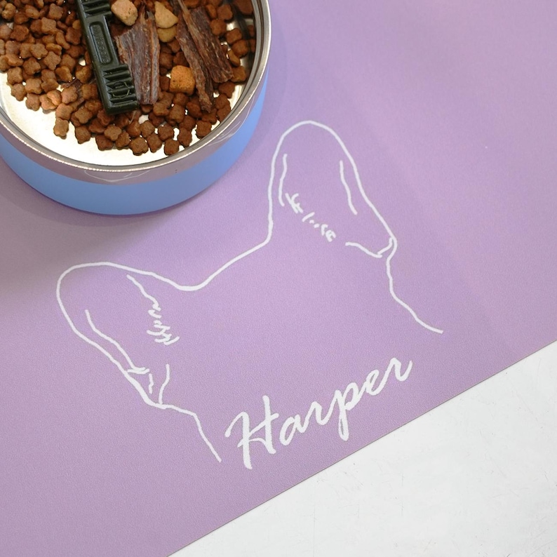 May include: A light purple pet food mat with a white outline of a dog's head and the name "Harper" in cursive. A blue and silver pet food bowl is partially visible, filled with dry food and treats.
