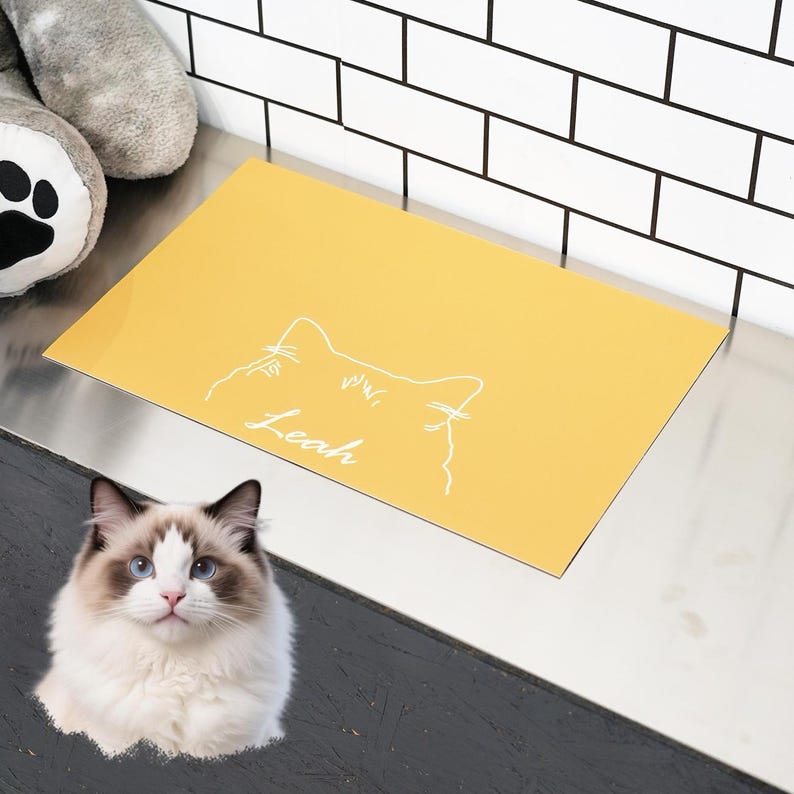 May include: A yellow pet placemat with a white cat outline and the name "Leah" in script. The placemat is rectangular and sits on a metallic surface. A fluffy cat with blue eyes is in the foreground.