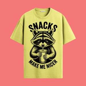 Może przedstawiać: Jasnożółta koszulka z czarno-białą ilustracją szopa pijaącego z kubka. Tekst "SNACKS MAKE ME NICER" jest wygięty nad i pod wizerunkiem szopa. Koszulka ma okrągły dekolt i krótkie rękawy.