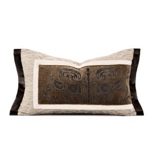 May include: Rectangular decorative pillow with a central brown panel featuring a black, stylized design. The panel is framed by a white border and set against a textured, light beige background. The pillow's ends are dark brown.