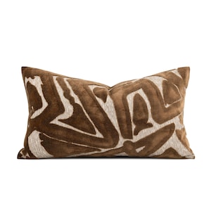 May include: A rectangular throw pillow featuring a textured, raised brown velvet pattern set against a light beige fabric background. The design consists of abstract, flowing shapes that cover the entire front surface of the pillow.