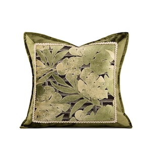 May include: A square throw pillow featuring a central panel with a green and light green tropical leaf pattern over a dark grid background. The panel is framed by a decorative cream-colored braided trim and a solid olive green velvet border.