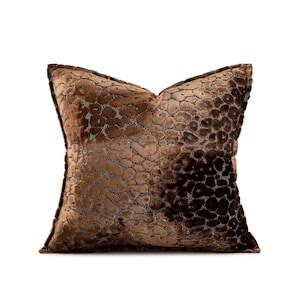 May include: A square decorative pillow with a brown, textured pattern. The pillow has a soft, velvet-like appearance and a dark brown border. The pattern is a repeating design of small, irregular shapes.
