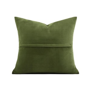 May include: Olive green velvet pillow with a rectangular shape. The pillow has a smooth texture and a solid color. A horizontal seam is visible on the back of the pillow. The pillow is designed for home decor.