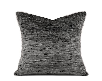 May include: Square decorative pillow with a textured, dark gray fabric. The pillow has a solid color with a subtle woven pattern. The edges are finished with a white trim, creating a contrast. The pillow is designed for home decor.