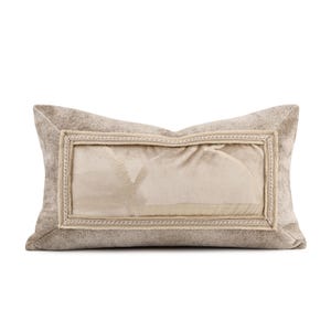 May include: A rectangular decorative pillow in neutral tones. The pillow features a central panel with a lighter shade, framed by a darker, textured fabric. A decorative border surrounds the central panel, adding a touch of elegance. The pillow is designed for home decor.