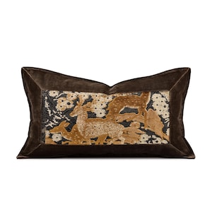 May include: A rectangular brown velvet pillow with a detailed embroidered scene of deer in a forest. The central panel features deer in shades of gold and brown against a black background with white floral accents. The pillow has a dark brown velvet border.