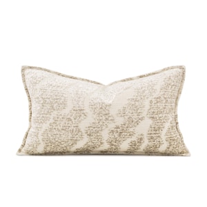 May include: A rectangular decorative pillow with a neutral color palette. The pillow features a textured, abstract pattern in shades of beige and light brown. The edges are finished with a subtle, darker trim, adding a touch of definition to the design.
