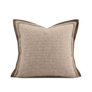 May include: A square decorative pillow with a textured, light beige center and a dark brown border. The pillow has a woven appearance and a soft, inviting aesthetic. The neutral color palette makes it suitable for various interior design styles.