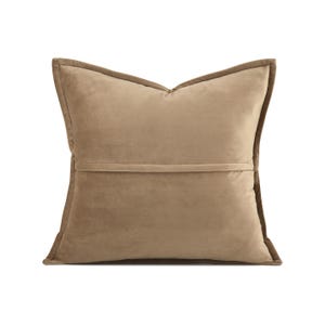 May include: A square, tan cushion with a soft, velvety texture. The cushion has a darker brown trim around the edges and a horizontal seam across the back. The cushion is designed for home decor.