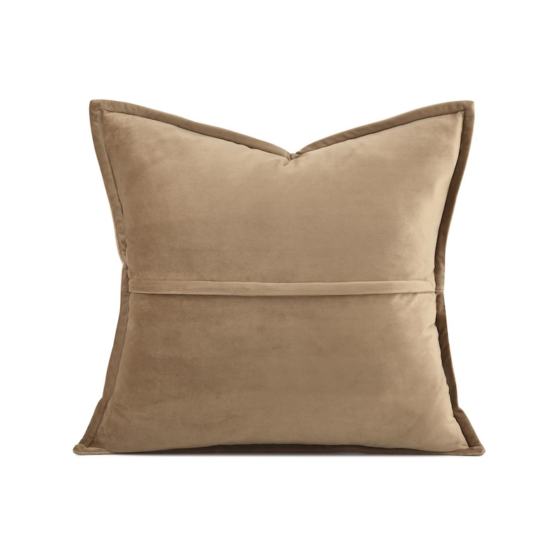 May include: A square, tan cushion with a soft, velvety texture. The cushion has a darker brown trim around the edges and a horizontal seam across the back. The cushion is designed for home decor.