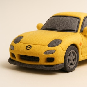 Op de afbeelding: Een gele speelgoedauto met zwarte accenten, waaronder de wielen, koplampen en raamkozijnen. De auto heeft een spoiler en zijspiegels. Het Mazda-logo is zichtbaar aan de voorkant.