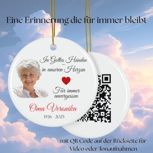 Pode incluir: Enfeite redondo de cerâmica branca com foto de mulher, coração vermelho e texto. O enfeite tem um fio dourado para pendurar. O texto inclui "Nas mãos de Deus, em nossos corações, para sempre inesquecível, Oma Veronika 1936-2025". Um código QR está na parte de trás.