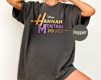 Peut inclure: T-shirt gris foncé avec l'inscription "Hannah Montana 20th Anniversary Special" en or, violet et blanc. Le logo Disney est au-dessus du texte. Le t-shirt a des manches courtes et un col en V.
