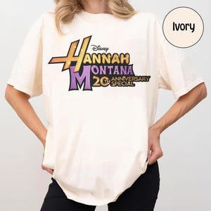 May include: Ivory-colored t-shirt featuring the Hannah Montana logo in gold, purple, and black. The design includes the text "Disney Hannah Montana 20th Anniversary Special". The shirt has short sleeves and a relaxed fit.