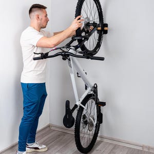 Pode incluir: Uma bicicleta de montanha branca montada numa prateleira de parede de madeira. A bicicleta tem pneus pretos e um assento preto. A prateleira está fixada a uma parede branca. Uma pessoa está a segurar a bicicleta.