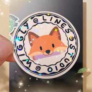 Peut inclure: Un autocollant rond avec un renard de dessin animé orange et un fond blanc. L'autocollant porte le texte "Wiggly Lines Studio" autour du renard. L'autocollant a un effet holographique et scintillant.