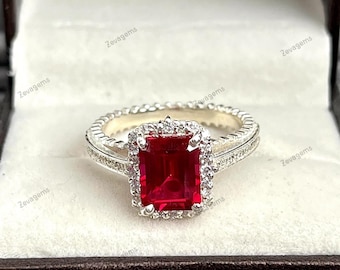 Op de afbeelding: Een zilverkleurige ring met een grote, rechthoekige, smaragdgeslepen rode edelsteen in het midden. De centrale steen is omringd door een halo van kleine, heldere, rondgeslepen steentjes. De ringband is voorzien van een textuur met kleine inkepingen.