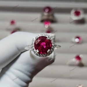 Könnte beinhalten: Ein silberfarbener Ring mit einem großen, runden, facettierten tiefroten Edelstein in der Mitte, umgeben von einem Kranz aus kleinen, klaren, funkelnden Steinen. Die Ringschiene ist schmal und ebenfalls mit kleinen, klaren Steinen besetzt.