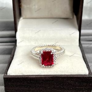 Op de afbeelding: Een zilveren ring met een grote, rechthoekige, rode edelsteen in het midden, omringd door kleinere witte stenen. De ring wordt gepresenteerd in een doos met een witte binnenkant.