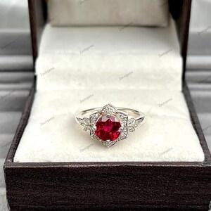 May include: A silver ring with a large, round, deep red gemstone at the center, surrounded by smaller clear stones and floral accents. The ring is presented in a dark brown jewelry box with a cream-colored interior.