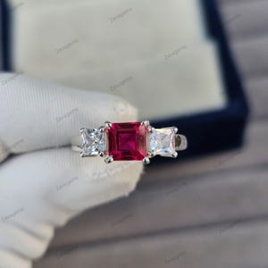 Könnte beinhalten: Ein silberner Ring mit einem großen, quadratischen, tiefroten Edelstein, flankiert von zwei kleineren, quadratischen, klaren Edelsteinen. Der Ring ist vor weißem Hintergrund zu sehen, mit dem Markennamen "Zevagems".