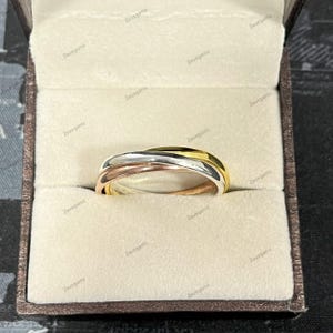 Op de afbeelding: Een driekleurige ring met verweven banden van goud, zilver en roségoud. De ring wordt gepresenteerd in een beige sieradendoosje met een bruine buitenkant. Het ontwerp van de ring is een moderne interpretatie van een klassieke stijl.