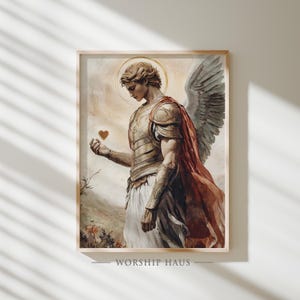 May include: A framed print featuring a detailed illustration of an angel. The angel, with a golden halo, wears armor and a red cape, holding a heart. The artwork has a soft, painterly style with a warm color palette. The text "WORSHIP HAUS" is at the bottom.