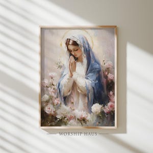 Puede incluir: Pintura enmarcada de una mujer con velo azul, con las manos juntas en oración, rodeada de rosas rosas y blancas. La obra de arte tiene un estilo suave y pictórico con un halo dorado. El texto "WORSHIP HAUS" está en la parte inferior.