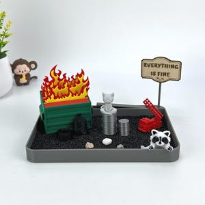 May include: A miniature diorama featuring a green dumpster engulfed in flames, with a sign that reads "EVERYTHING IS FINE." Other elements include a cat statue, a red crane, and a raccoon figurine. The scene is set on a gray tray.