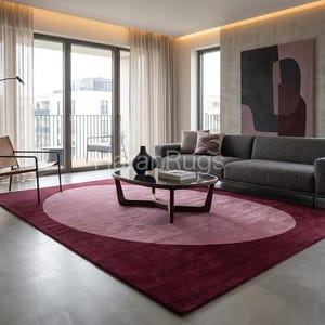 Könnte beinhalten: Ein modernes Wohnzimmer mit einem großen, ovalen Teppich in tiefroten und rosa Farbtönen. Ein dunkelgraues Sofa, ein Couchtisch und ein Ledersessel ergänzen das Design. Große Fenster und neutrale Vorhänge sorgen für natürliches Licht. Der Teppich misst ca. 300 cm x 200 cm.