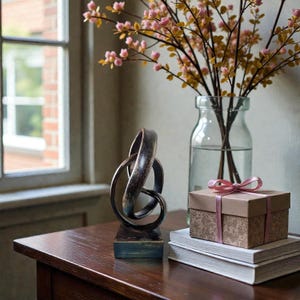 May include: A decorative bronze-colored sculpture with an abstract design sits on a wooden base. A glass vase holds branches with pink flowers. A small gift box with a pink ribbon rests on a stack of books.