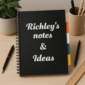 May include: A black spiral-bound notebook with the words "Richley's notes & Ideas" in white script. The notebook has colourful divider tabs. A pen, pencil, eraser, and ruler are on the wooden desk.